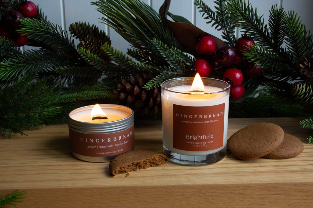 Holiday Collection from Brightfield, Gingerbread candles burning with a gingerbread cookie beside them and holiday greenery, and Christmas scented candles, holiday candles, wood wick candles, Gingerbread candle, best Christmas candles