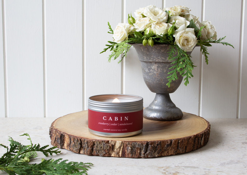 A cabin travel candle from Brightfield sits on a wood log with a vase of white roses and cedar branches beside it. Cosy aesthetic, fall candle, winter candle, cranberry candle, cedar candle, sandalwood candle, candles for sale 