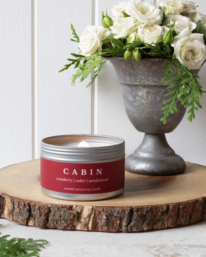 Cabin travel candle from Brightfield sits on a wooden log base in front of a vase of white roses and cedar branches. Gifts under $30, birthday gifts, candles under $30, cute gift ideas, wood wick candles for sale in Canada