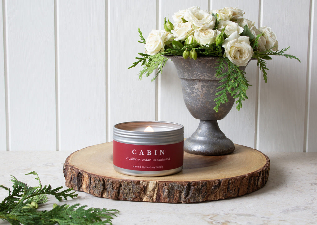 Cabin travel candle from Brightfield sits on a wooden log base in front of a vase of white roses and cedar branches. Gifts under $30, birthday gifts, candles under $30, cute gift ideas, wood wick candles for sale in Canada