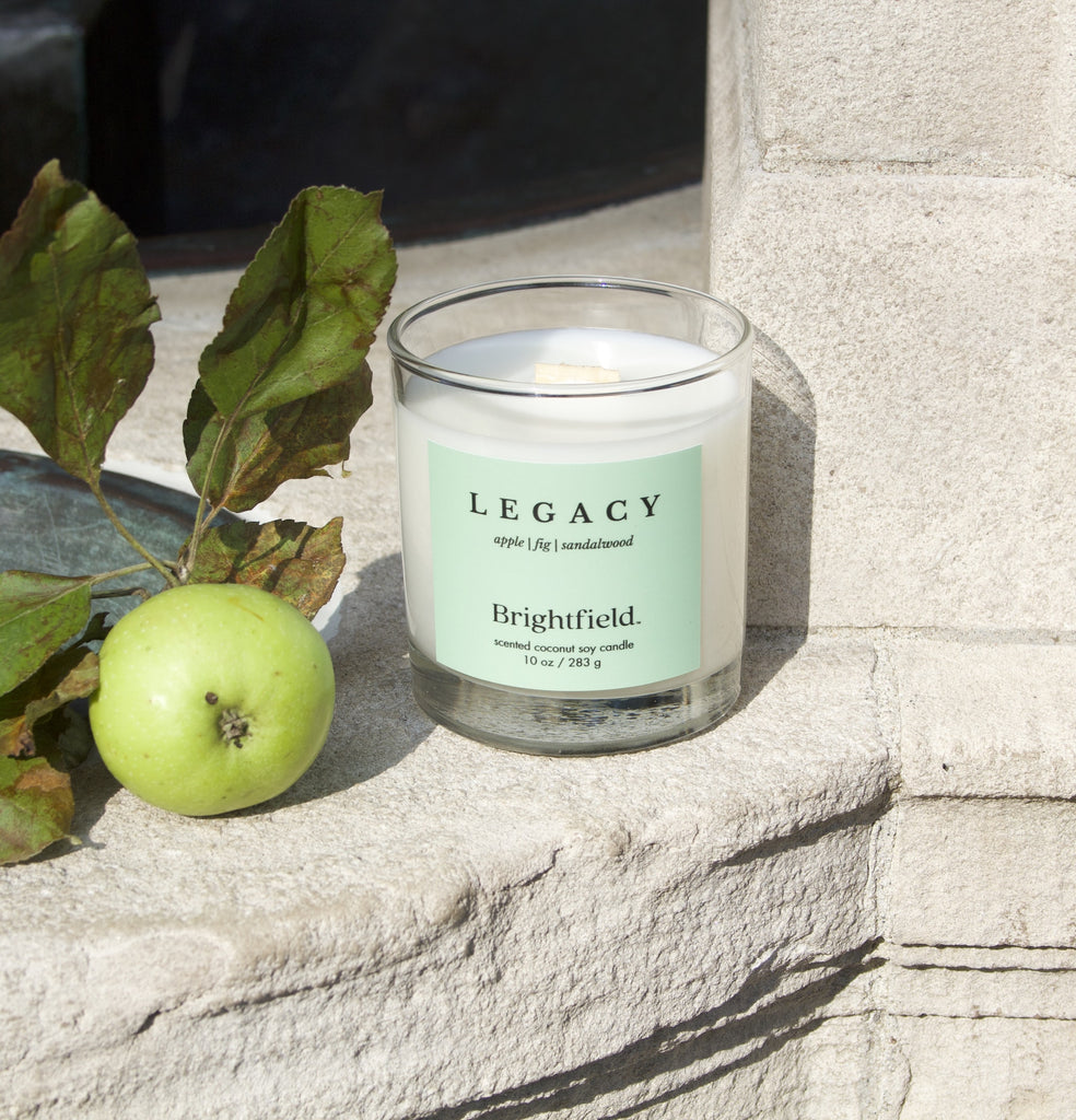 Wood wick candle in glass jar sits on a stone monument with apples beside it. The scent is Legacy from Brightfield, a Toronto candle shop