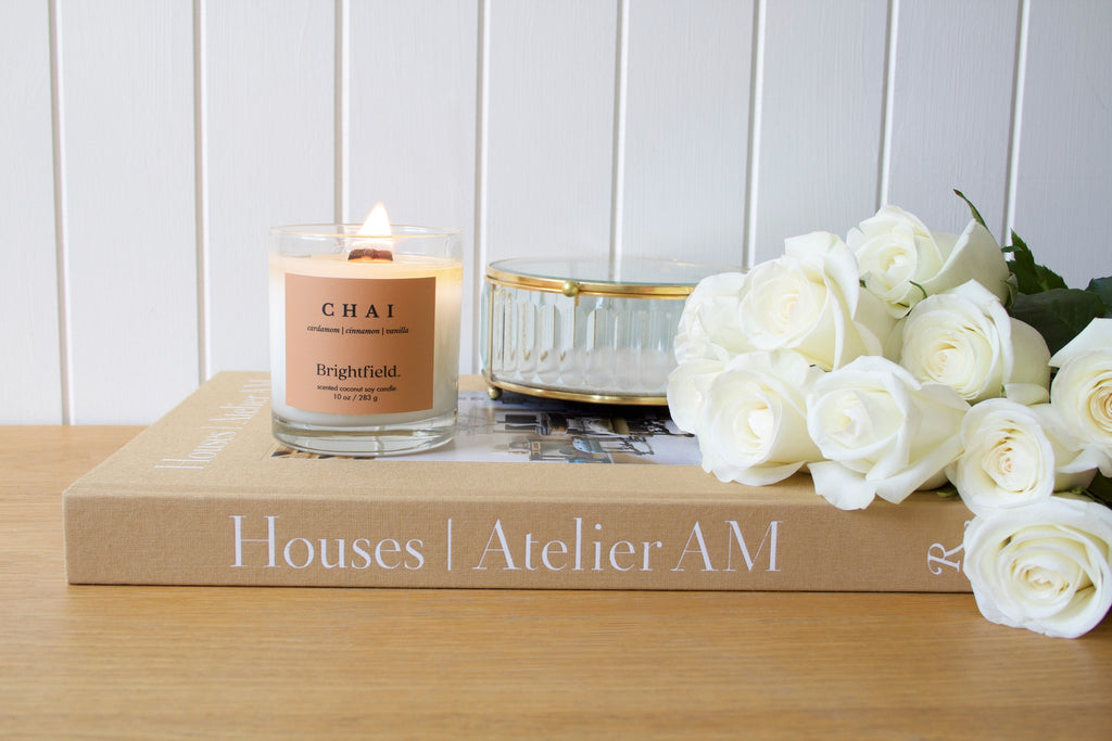 Chai candle from Brightfield sitting on a coffee table book with white roses beside it, Houses Atelier AM book, and a glass jewellery box. The Chai scented candle from Brightfield has notes of cardamom, cinnamon and vanilla